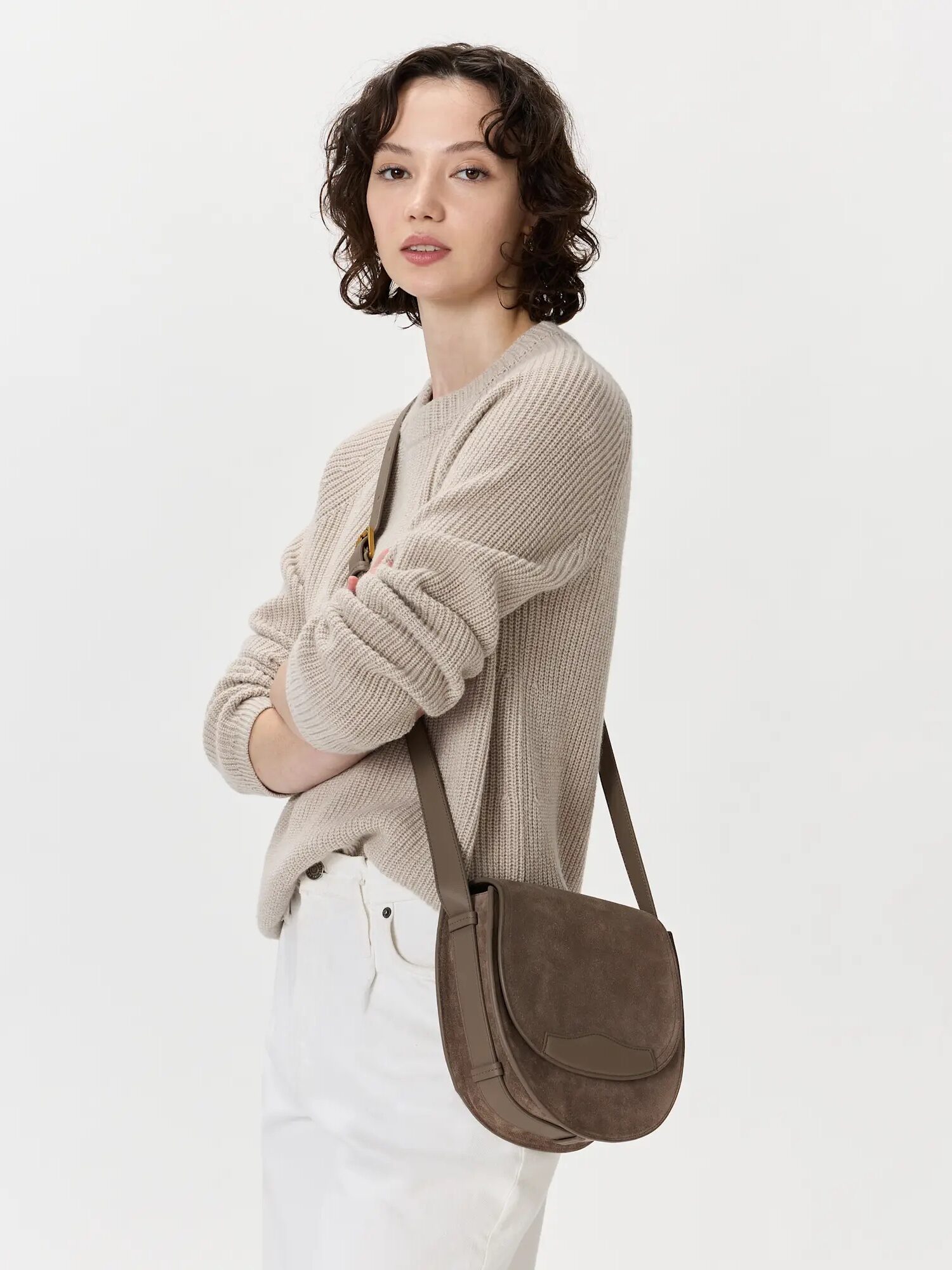 A woman wearing a beige sweater and white pants stands with arms crossed, carrying a taupe shoulder bag against a plain light background.