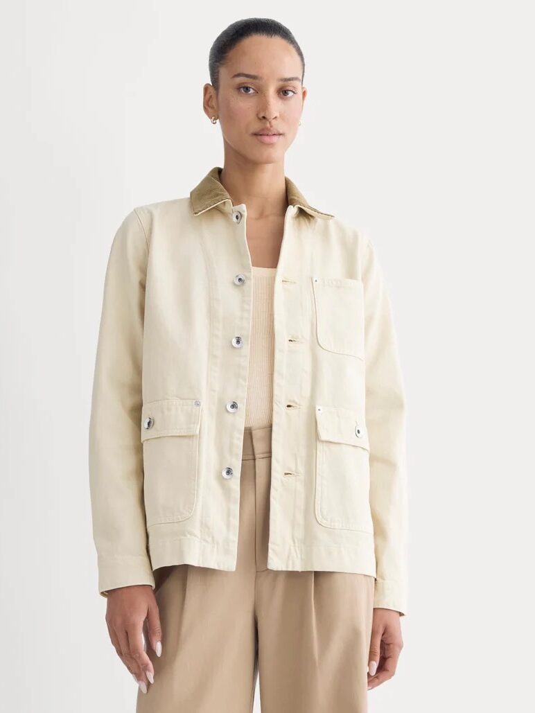 A woman stands facing forward, wearing a beige utility jacket with front pockets and tan pants, against a plain light background.