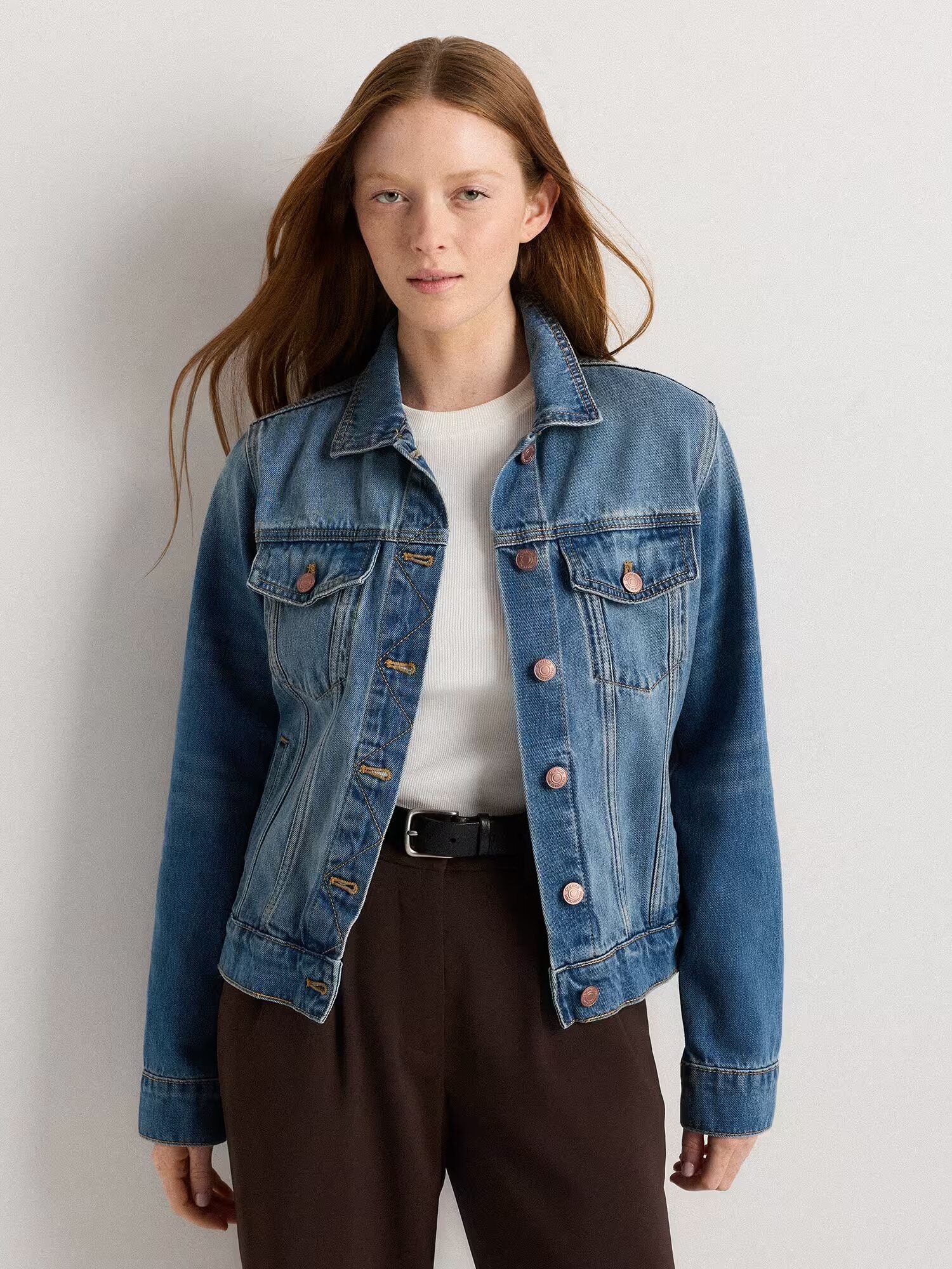 A person with long brown hair wears a blue denim jacket over a white shirt and brown pants, standing against a plain light background.