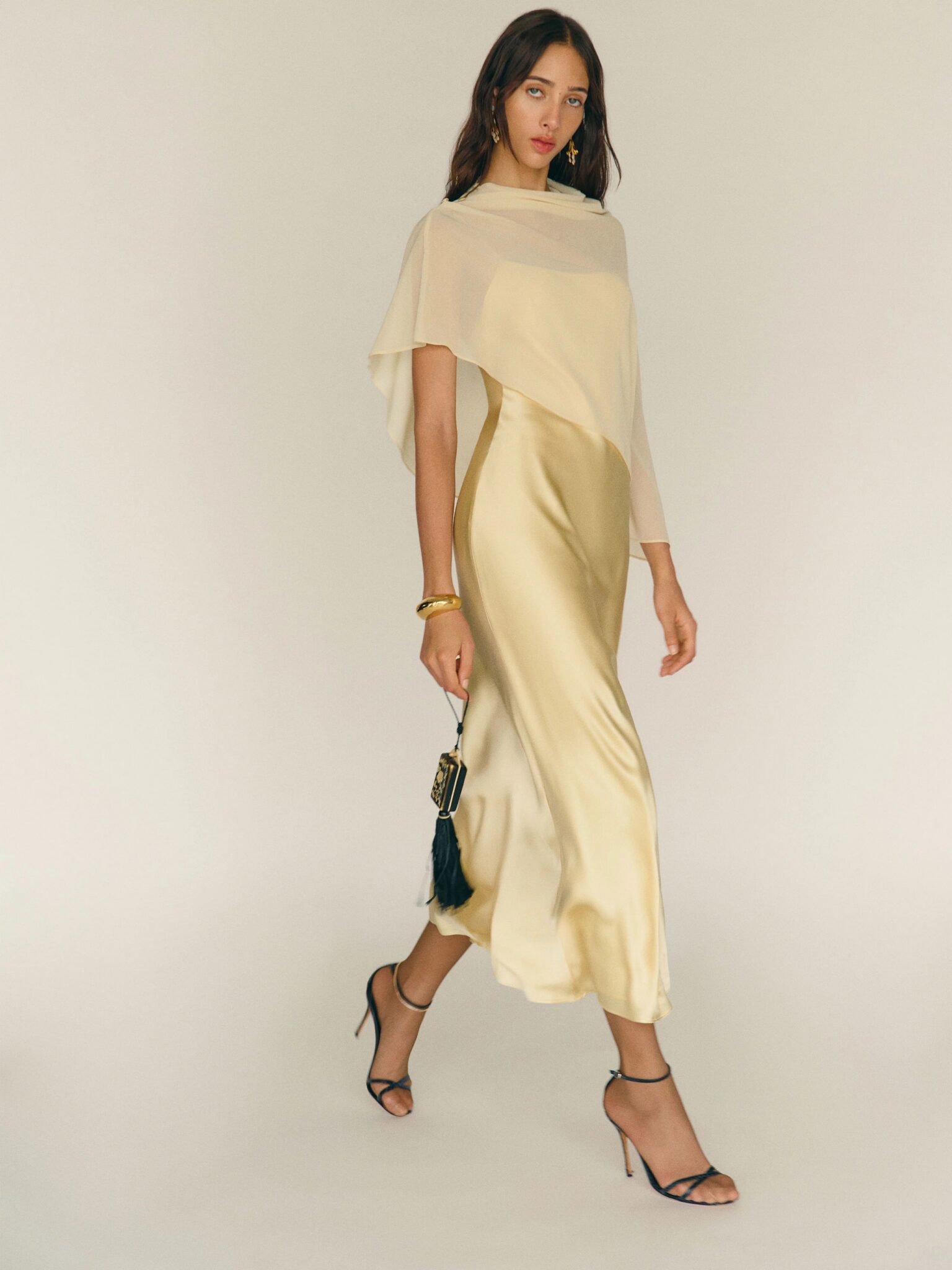 A woman wearing a flowing pale yellow dress and black high heels walks forward, holding a small black purse against a plain light background.
