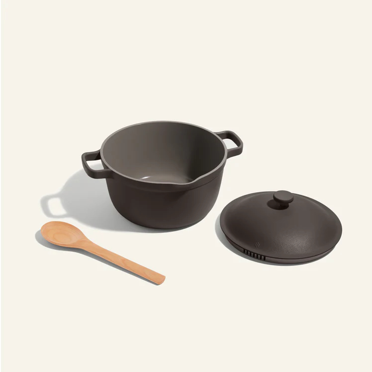 A brown, round Dutch oven with side handles, a matching lid placed to the right, and a wooden spoon positioned on the left, all set against a plain light background.