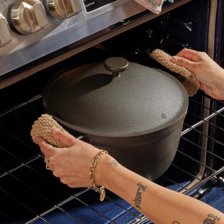 A person uses oven mitts to remove a lidded cast iron pot from an oven.
