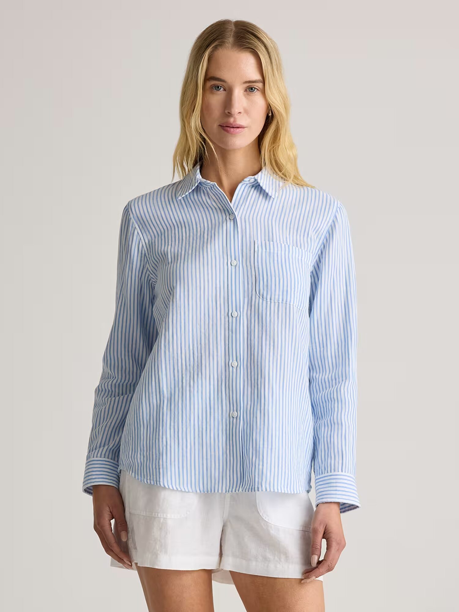 A person wearing a light blue and white striped button-up shirt and white shorts stands facing forward against a plain background.