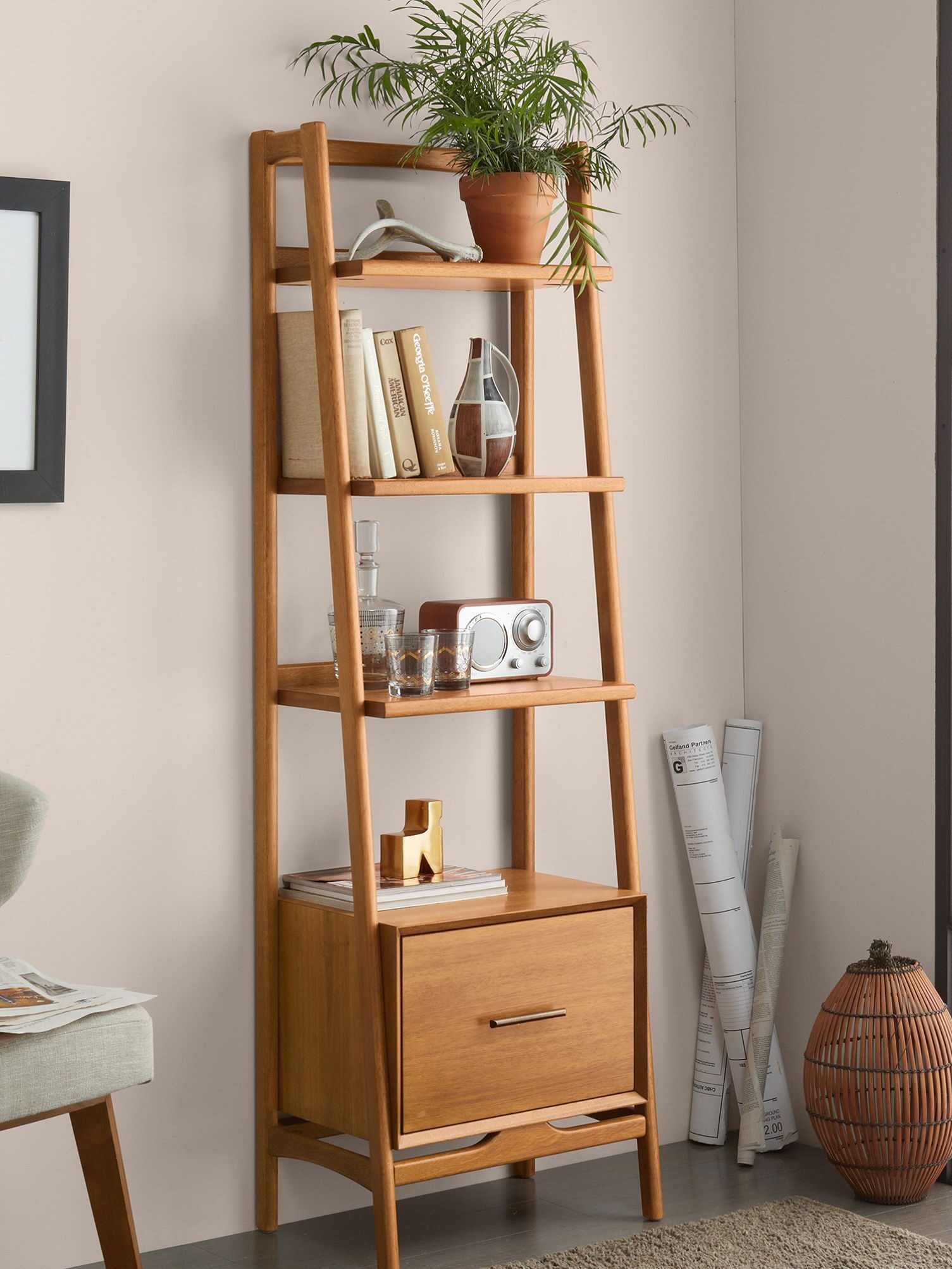 A wooden ladder bookshelf with books, a potted plant, glassware, a radio, and decor items. Rolled papers and a basket are beside it. A framed artwork hangs on the wall.