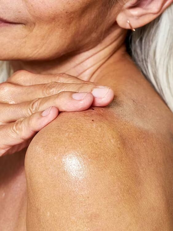 Close-up of an older woman with gray hair applying lotion to her bare shoulder, showing moisturized skin and minimal jewelry.