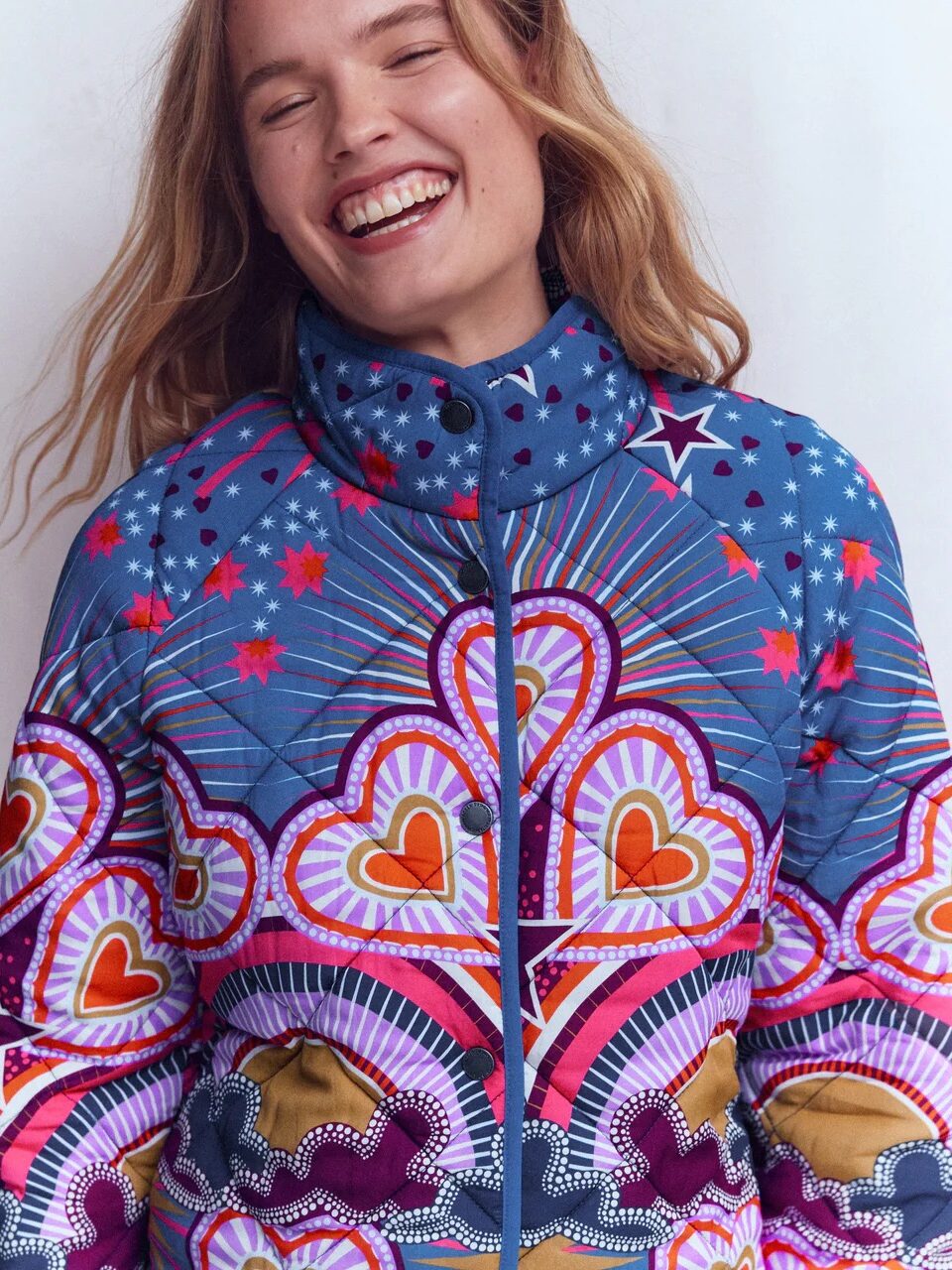 A woman with long hair smiles, wearing a colorful, patterned jacket featuring hearts, stars, and geometric designs in blue, pink, red, and gold tones.