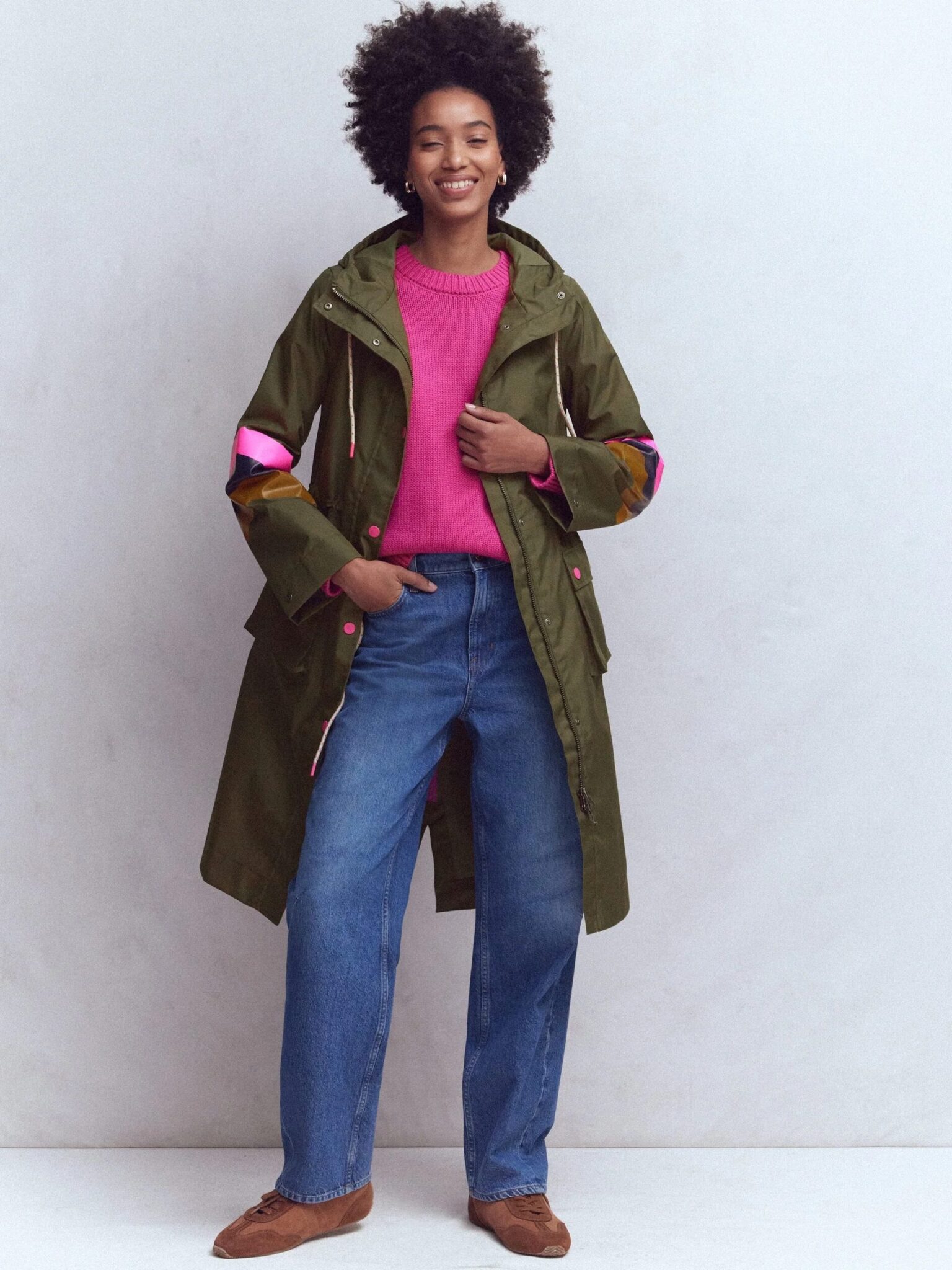 A person stands smiling, wearing a green parka with pink accents over a pink sweater, blue jeans, and brown shoes, against a plain light-colored background.