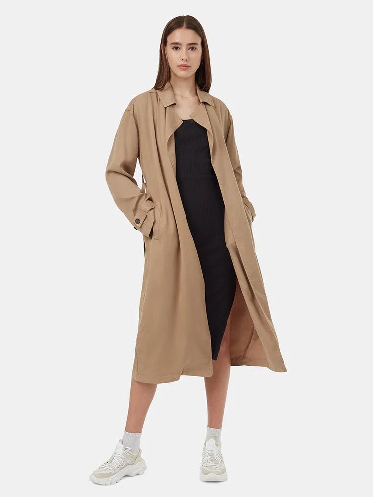 A woman stands wearing a long tan trench coat over a black dress with white socks and sneakers, facing forward against a plain white background.