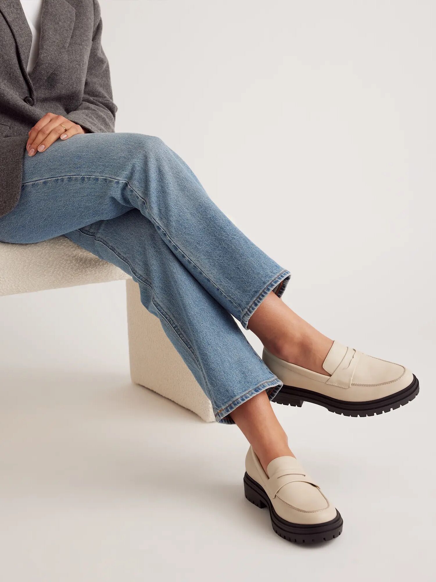 Person seated on a chair, wearing light blue jeans, a grey blazer, and cream-colored loafers with black soles.