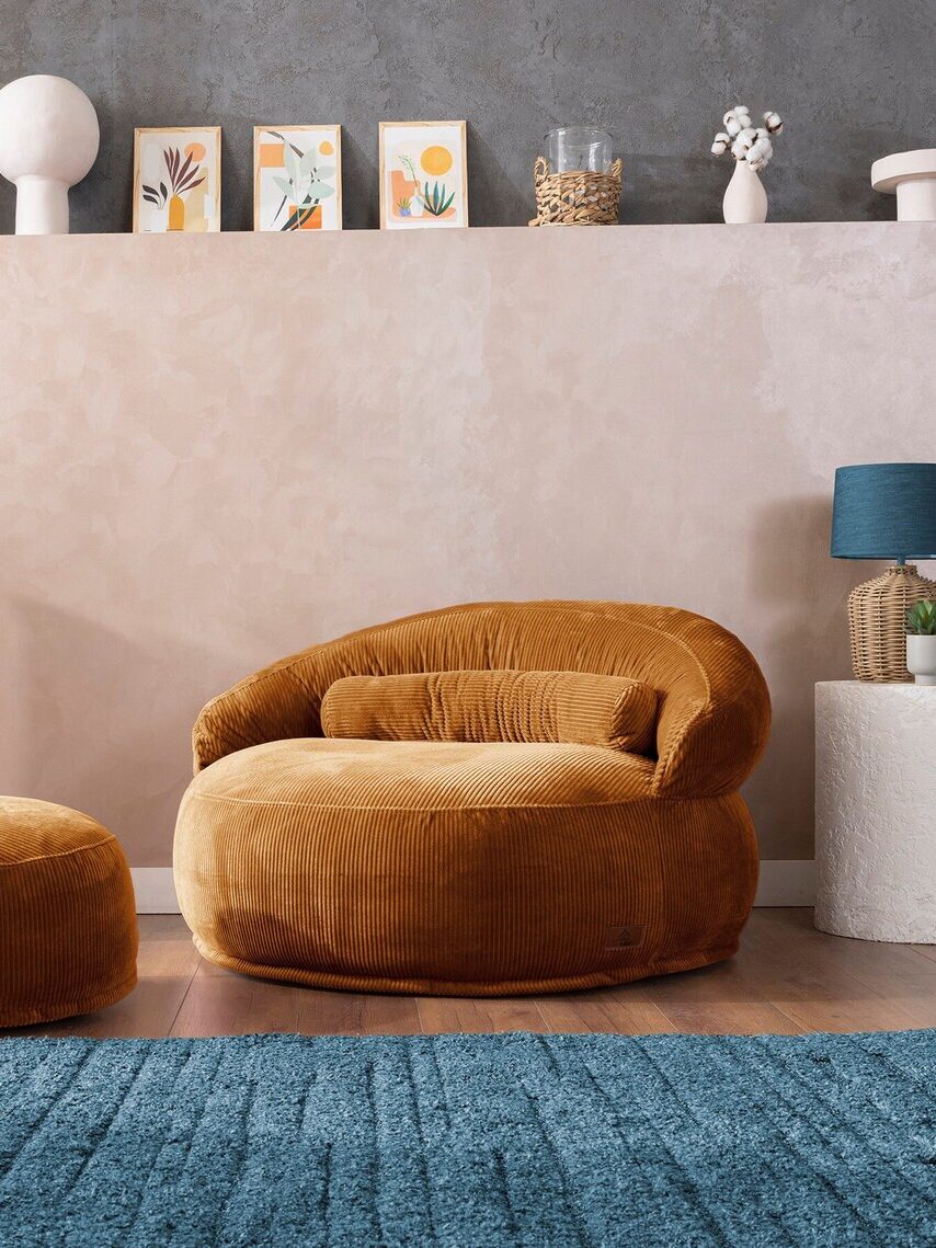 A mustard-colored corduroy lounge chair with a matching ottoman sits on a blue rug. Behind them, a shelf displays decorative objects, small framed art, and potted plants.