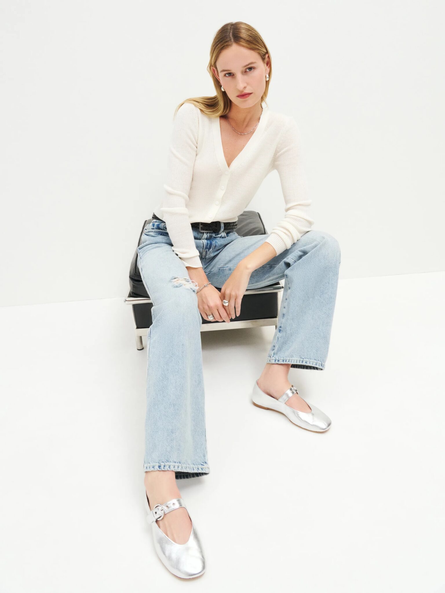 A woman sits on a black chair against a white background, wearing a white long-sleeve top, light blue ripped jeans, and silver flat shoes.
