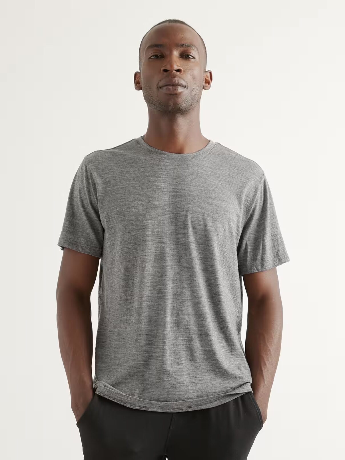 A man stands against a plain background wearing a heather gray short-sleeve t-shirt and black pants, with his hands in his pockets.