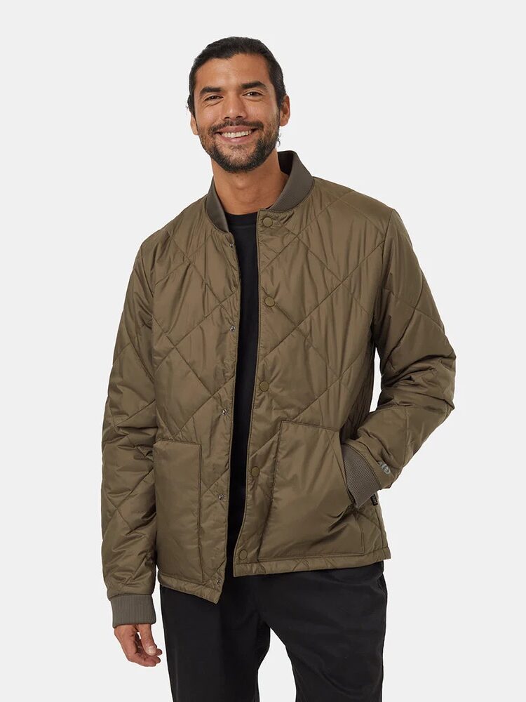 A man stands against a plain background wearing an olive green quilted jacket over a black shirt and black pants.