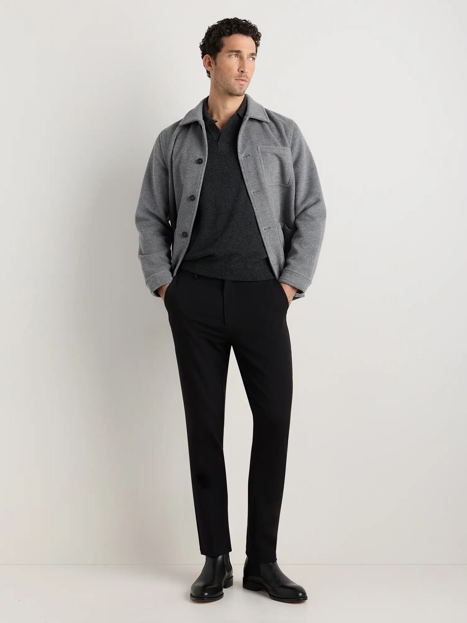 A man stands against a plain white wall wearing a grey jacket over a dark sweater, black pants, and black shoes.