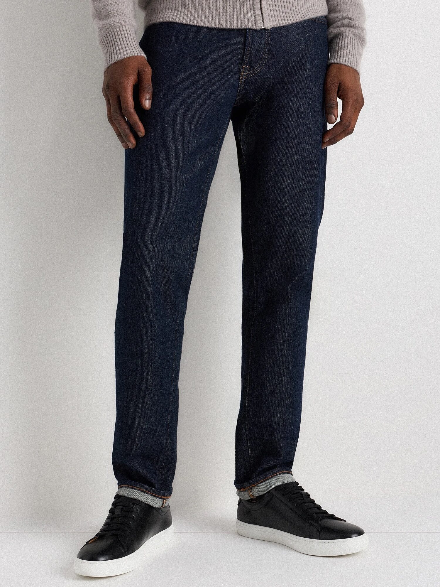 A person wearing a grey sweater, dark blue jeans, and black sneakers with white soles standing against a plain background.