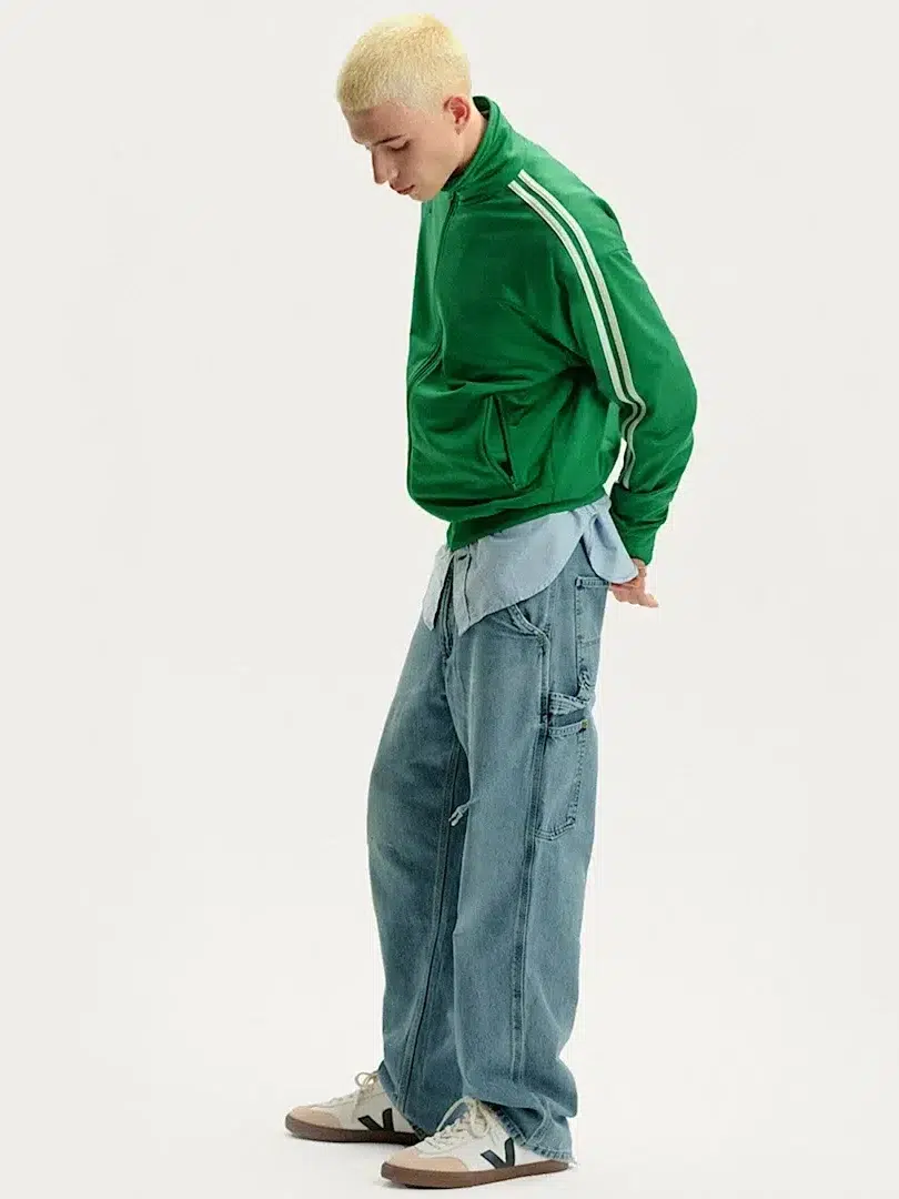 A person with short blond hair wears a green track jacket, blue shirt, baggy jeans, and white sneakers, standing against a plain light background.