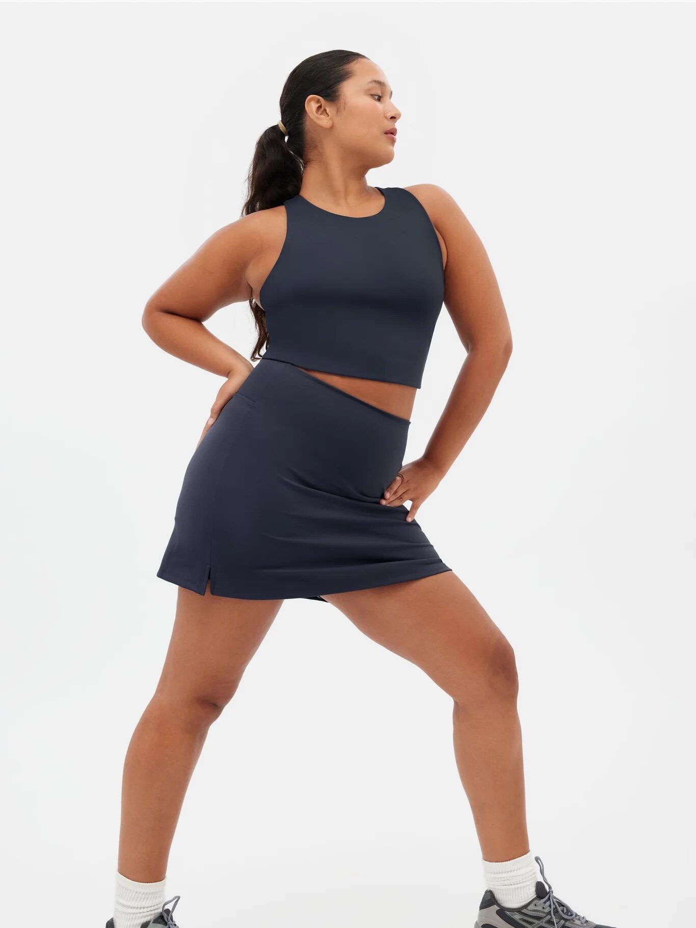 A person wearing a navy blue sleeveless crop top and matching skirt poses with one hand on their hip, dressed in gray sneakers and white socks against a plain background.
