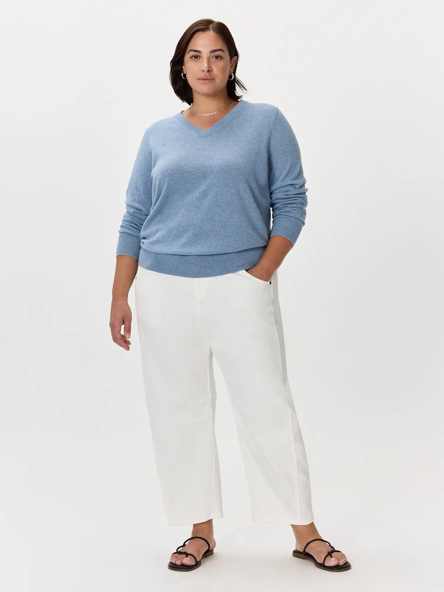 A person stands against a plain white background wearing a light blue V-neck sweater, white wide-leg pants, and black sandals.