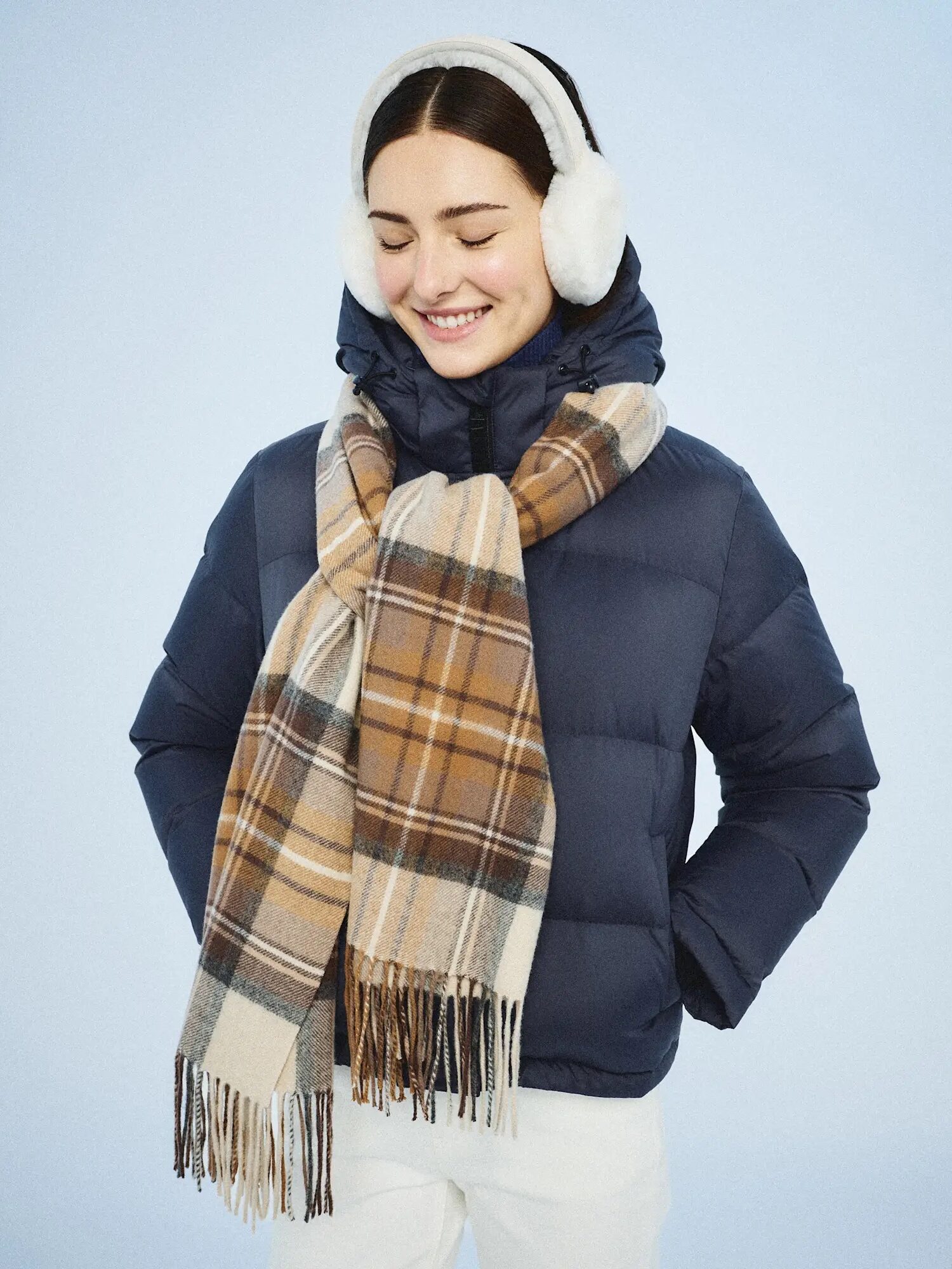 A person wearing a navy puffer jacket, plaid scarf, white earmuffs, and white pants stands against a light blue background.