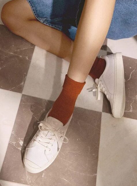 A person sits on a wicker chair with legs crossed, wearing a striped shirt, denim skirt, rust-colored socks, and white sneakers; close-up of the shoes shown on the right.