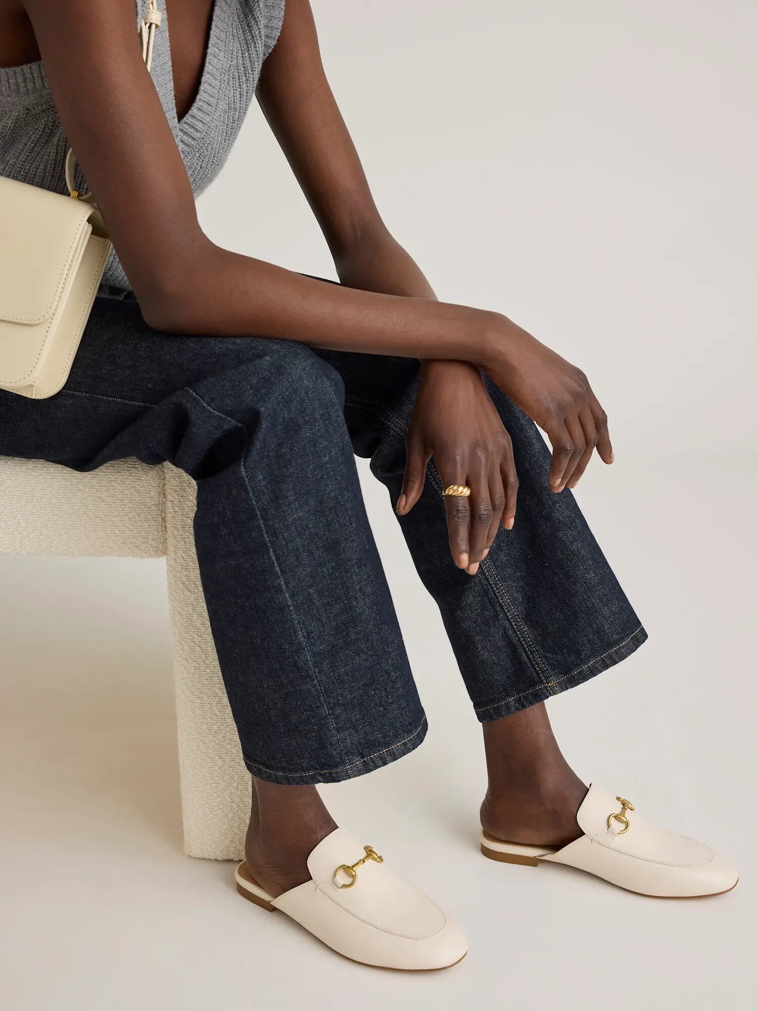 A person wearing dark jeans, a gray sleeveless top, cream-colored loafers with gold hardware, and a light beige bag sits on a textured white chair.