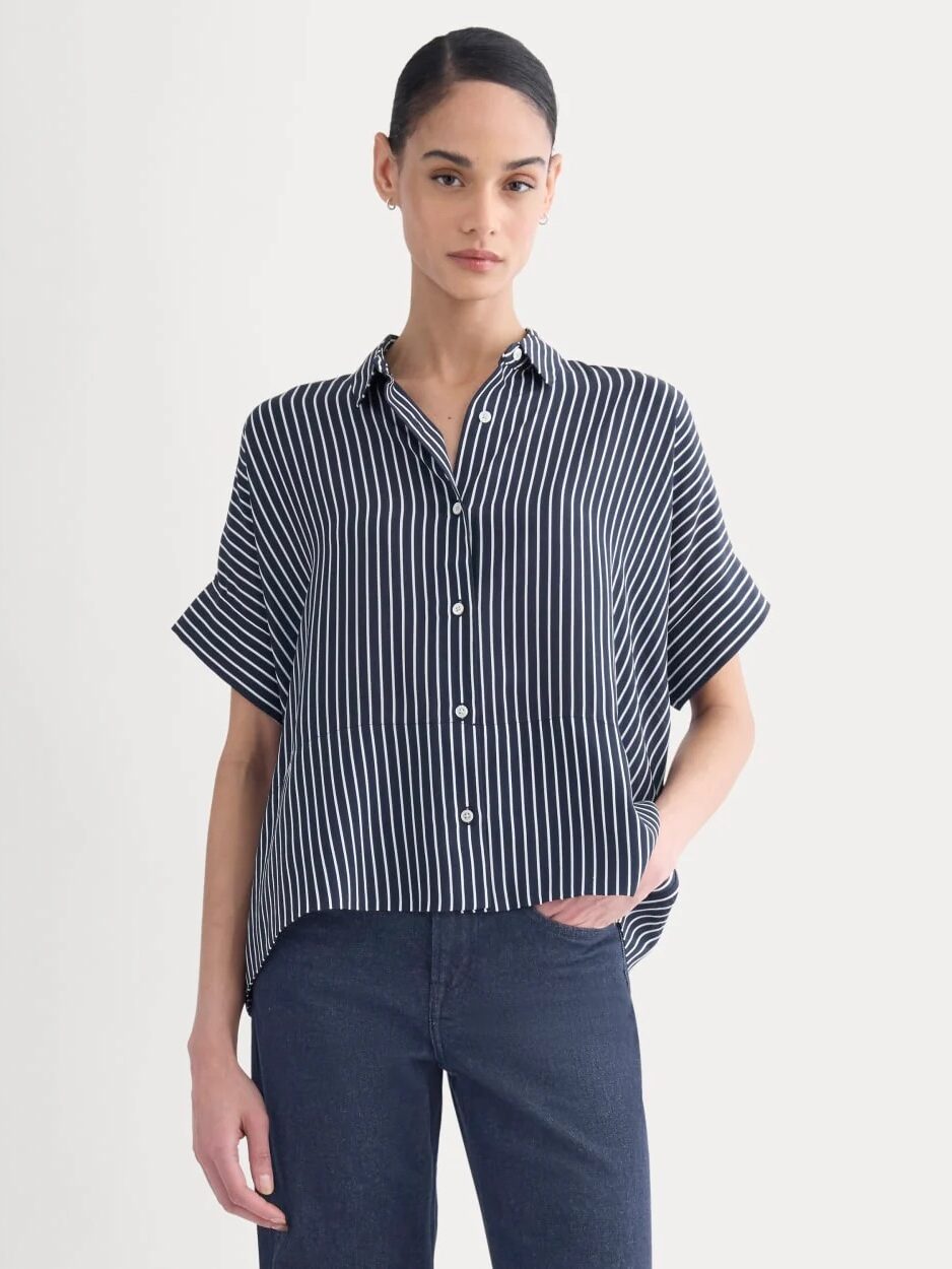 Woman wearing a short-sleeve, navy blue button-up shirt with white vertical stripes and dark jeans, standing against a plain light background.