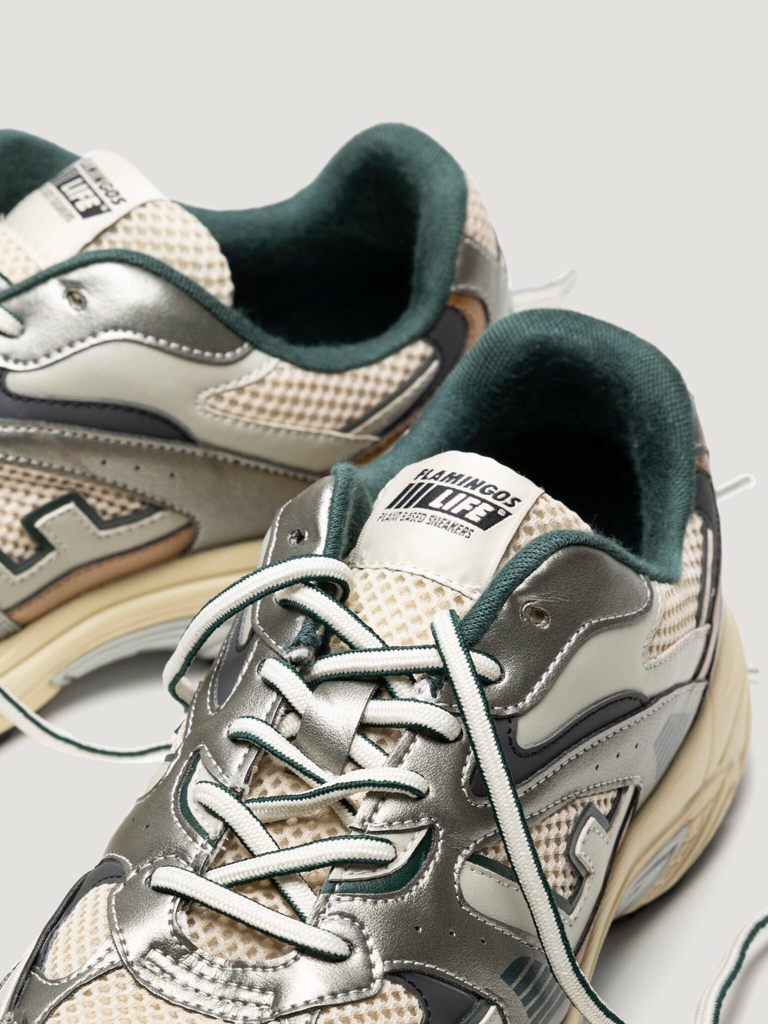 A close-up view of two metallic silver and beige sustainable sneakers with teal details and white laces on a light background.