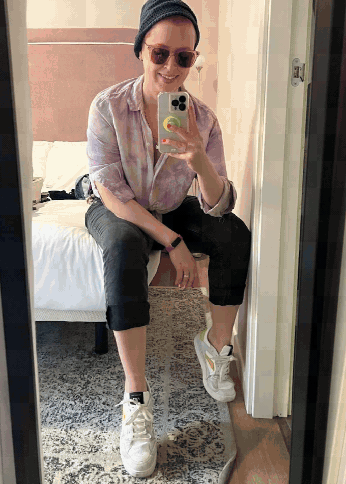 Person wearing sunglasses, a beanie, and casual clothes with sustainable sneakers takes a mirror selfie while sitting on the edge of a bed in a softly lit room.