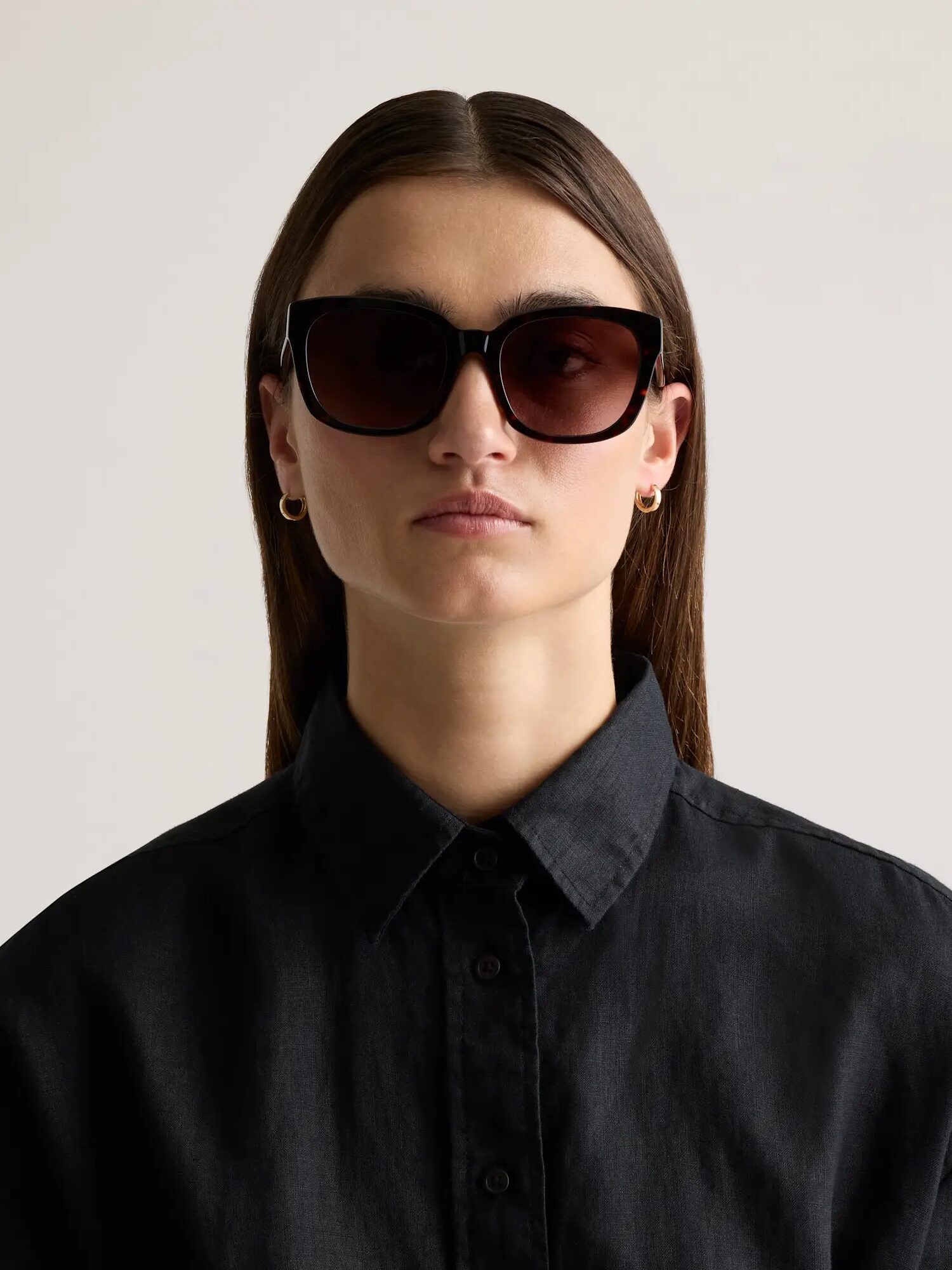 Person wearing black sunglasses, gold hoop earrings, and a black collared shirt, facing forward against a plain background.