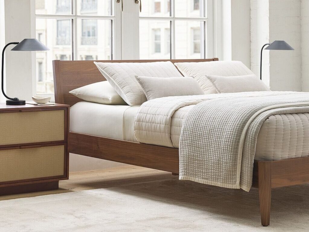 A modern bedroom featuring a wooden bed with an organic mattress, neutral bedding, a nightstand, two black lamps, a large window, and light-colored walls and carpet.