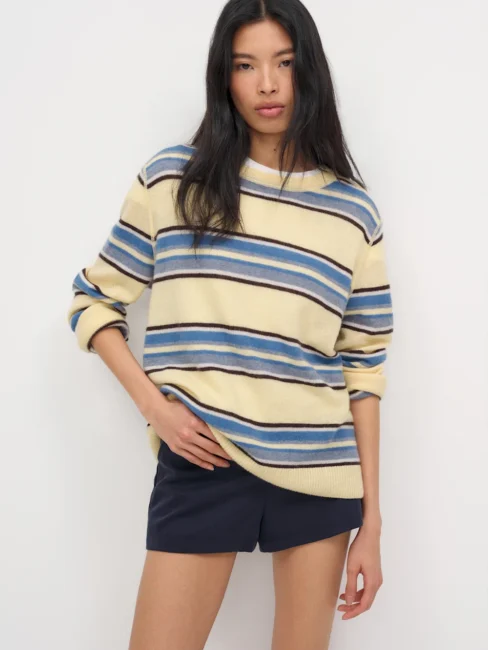 Person standing against a plain background, wearing a cream sweater with blue and beige horizontal stripes and dark shorts.