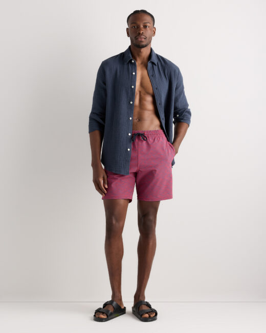 A man stands against a plain background wearing a navy blue button-up shirt, pink swim trunks, and black sandals.