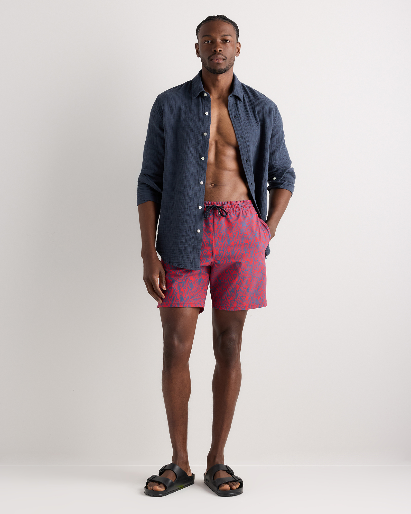 A man stands against a plain background wearing a navy blue button-up shirt, pink swim trunks, and black sandals.