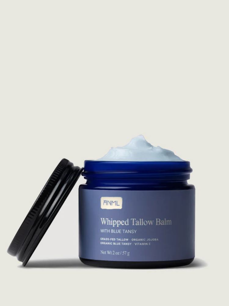 A blue jar of ANML Whipped Tallow Balm with Blue Tansy is open, showing light blue whipped balm inside. The label lists ingredients and the net weight as 2 oz / 57 g.