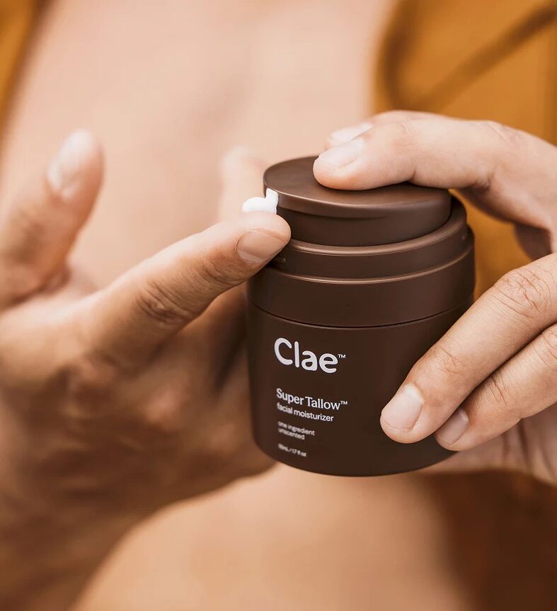 A person dispenses a small amount of Clae Super Tallow facial moisturizer onto their fingertip from a brown container.