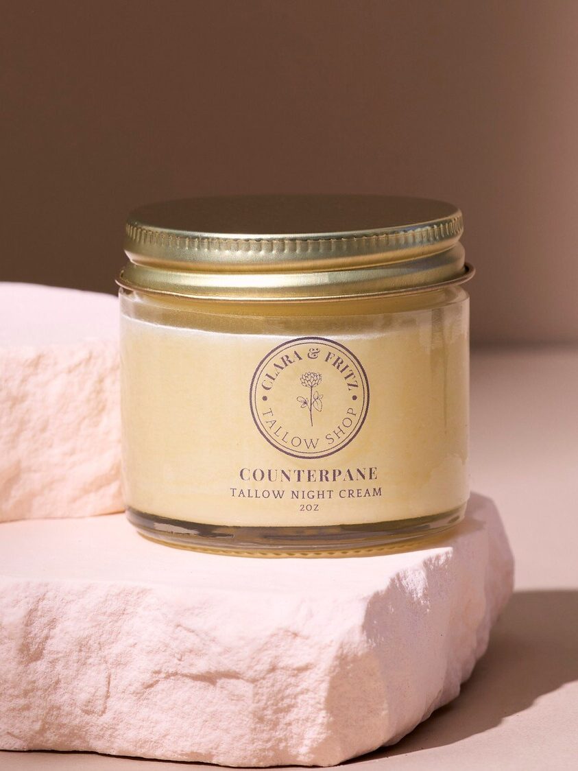 A glass jar of Counterpane Tallow Night Cream with a gold lid sits on a textured white stone surface against a neutral background.