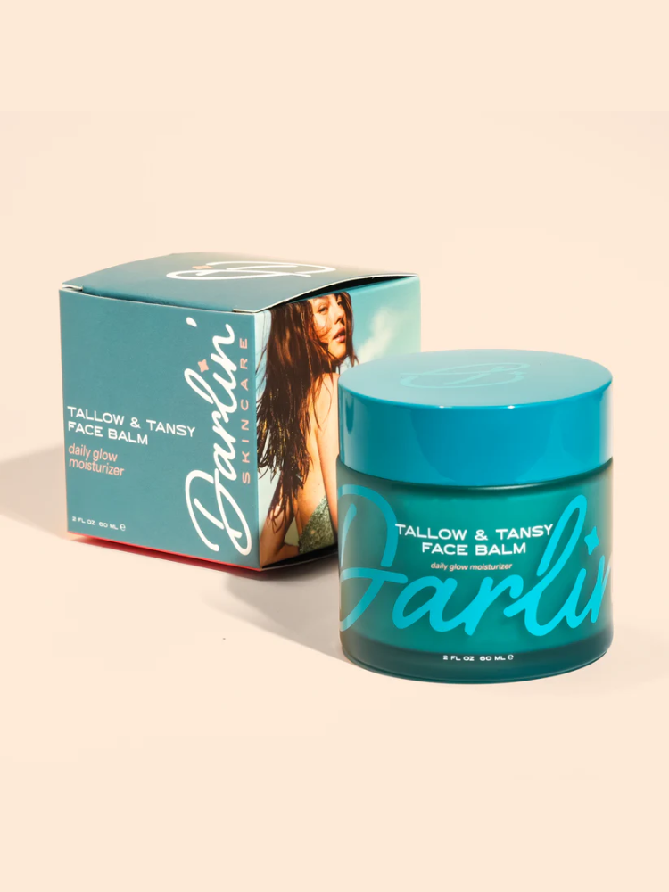 A teal jar labeled "Tallow & Tansy Face Balm" is displayed in front of its packaging, which features product details and an image of a woman.