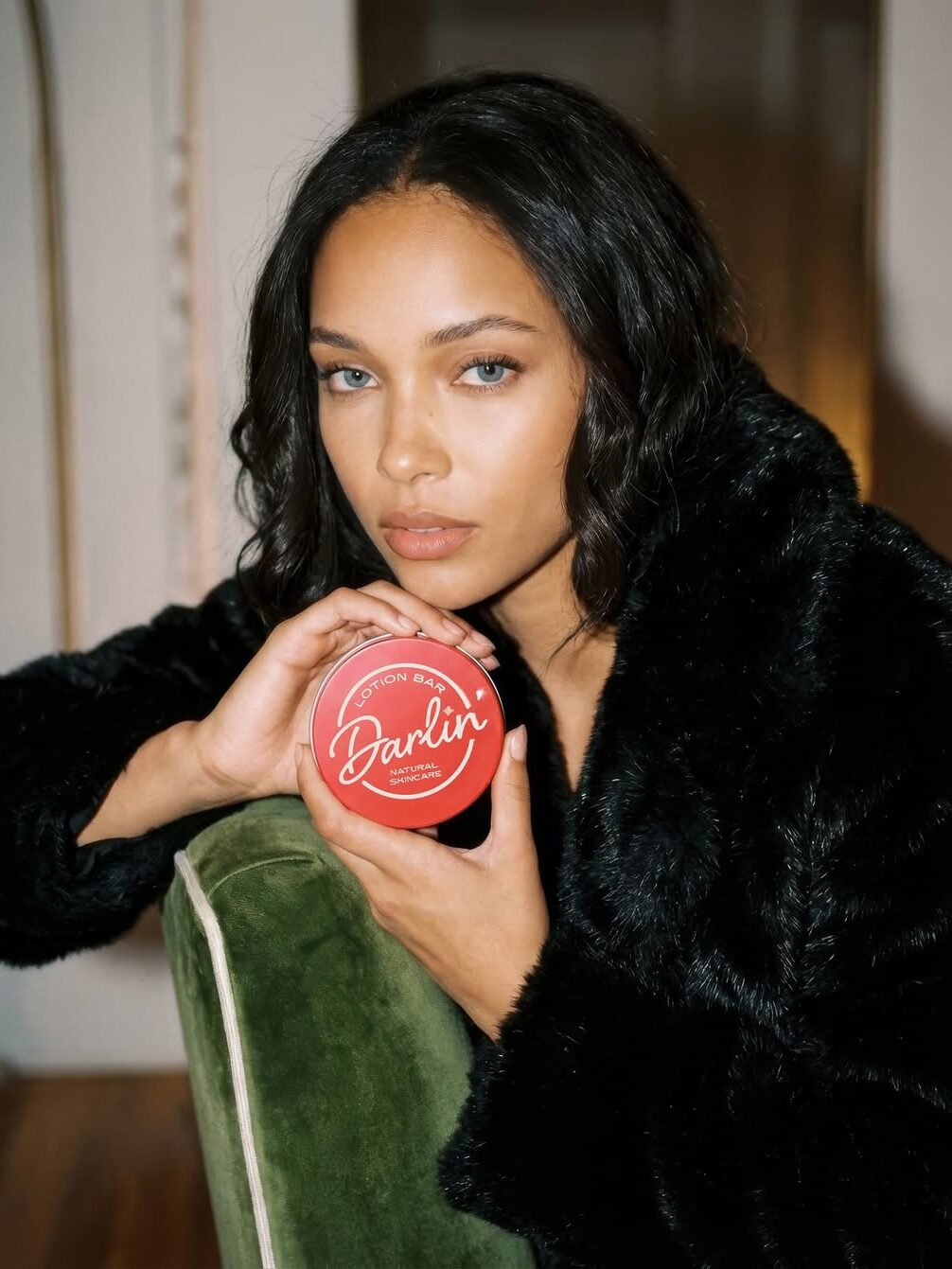 A woman with wavy hair in a black fur coat holds a round red container labeled "Darlin’" while sitting on a green chair.