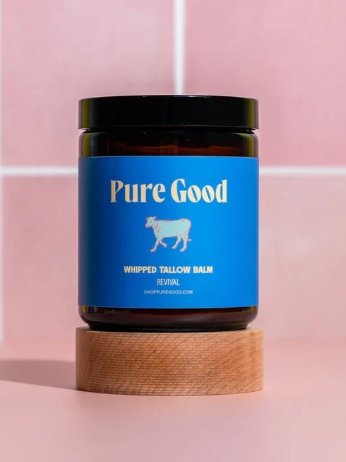 A jar labeled "Pure Good Whipped Tallow Balm" sits on a round wooden pedestal against a pink tiled background.