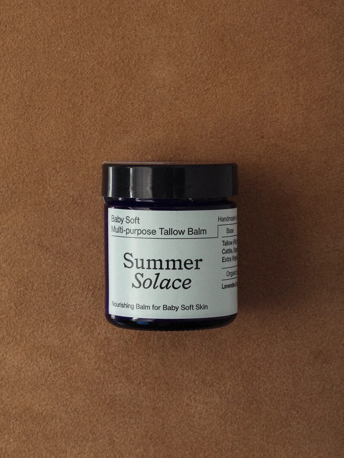 A dark jar labeled "Summer Solace Baby Soft Multi-Purpose Tallow Balm" sits on a brown surface. The label mentions it is handmade, organic, and for baby soft skin.