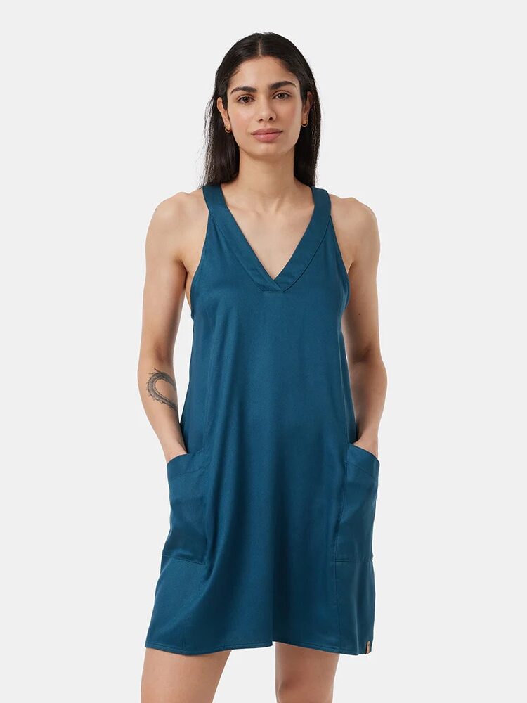 A woman stands facing forward wearing a sleeveless teal V-neck dress with pockets, against a plain white background.