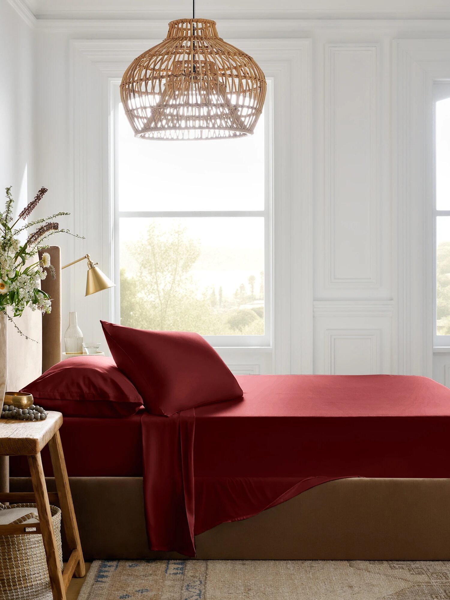 A neatly made bed with deep red sheets and pillows, placed next to a wooden nightstand with a lamp and flowers, under a woven pendant light in a bright, airy bedroom.
