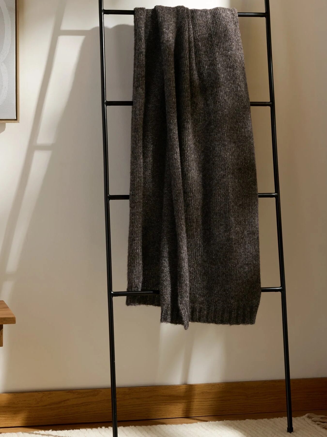 A dark gray knit blanket hangs on a black metal ladder in a sunlit corner next to a wooden table with a lamp, books, and a decorative box.
