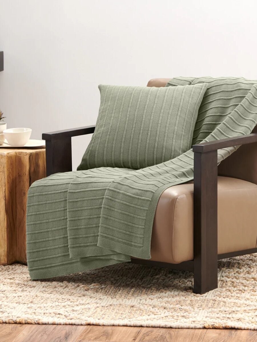 A tan leather armchair with a green ribbed throw blanket and matching pillow, next to a wooden side table with a cup and saucer, on a woven rug.