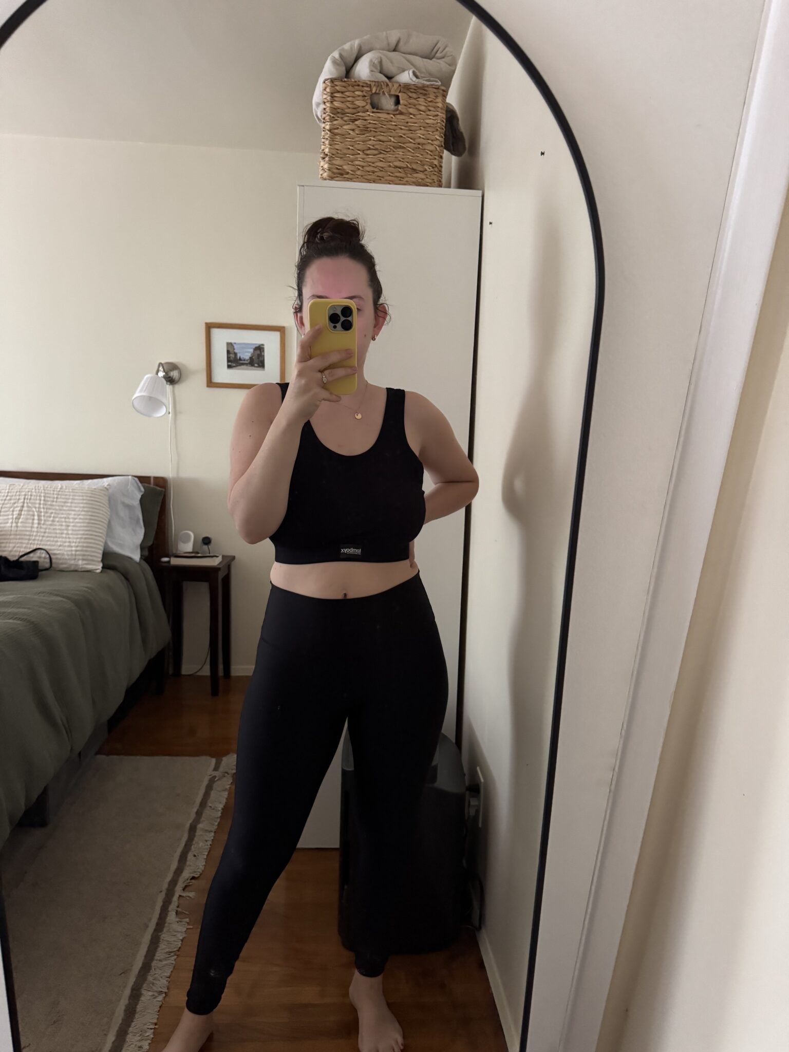 A person in black athletic wear takes a mirror selfie in a bedroom with a neatly made bed, a rug, and a basket on a shelf.