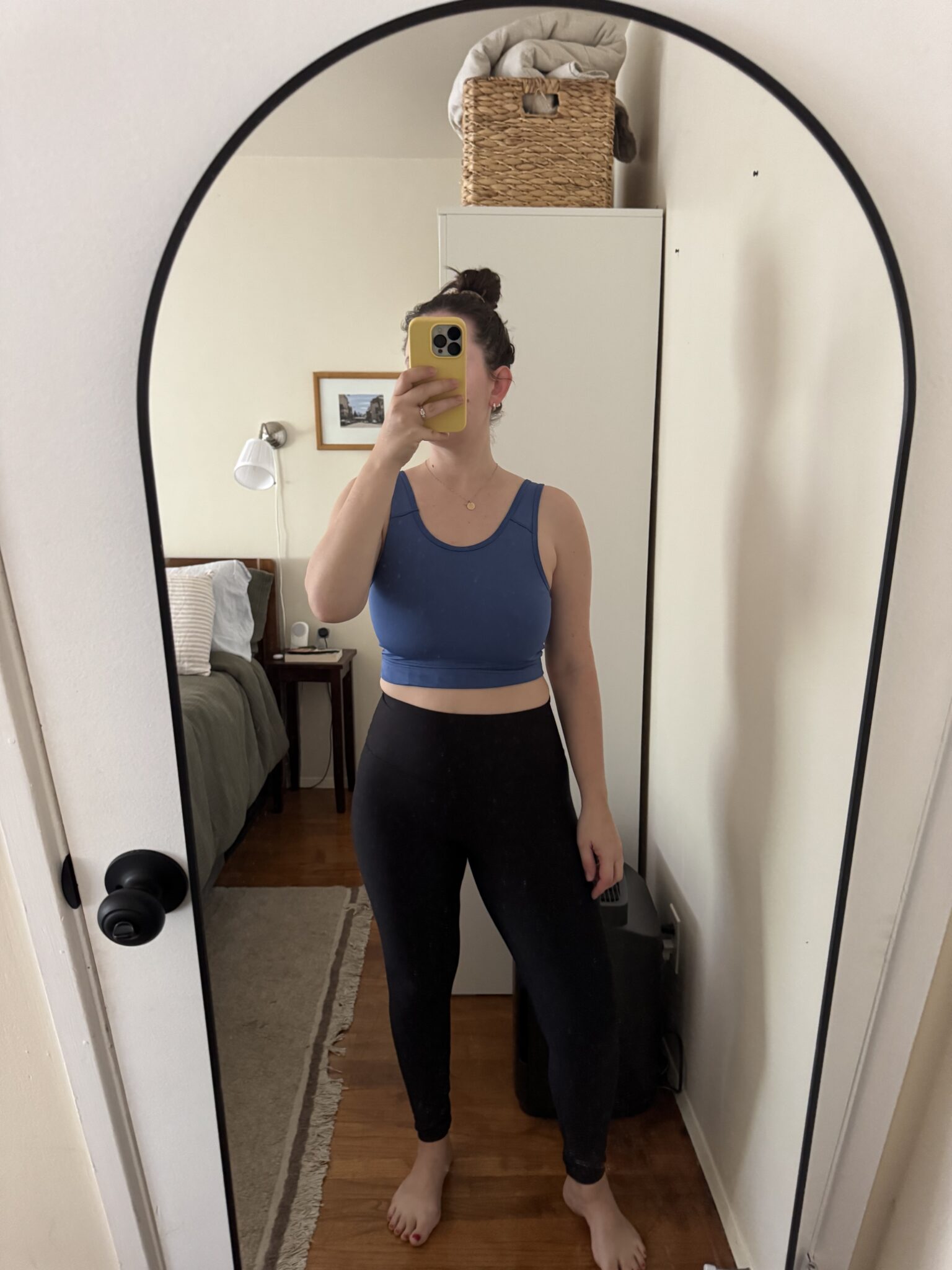 Person taking a mirror selfie in a bedroom, wearing a blue crop top and black leggings, with hair in a bun. A bed, nightstand, and basket are visible in the background.