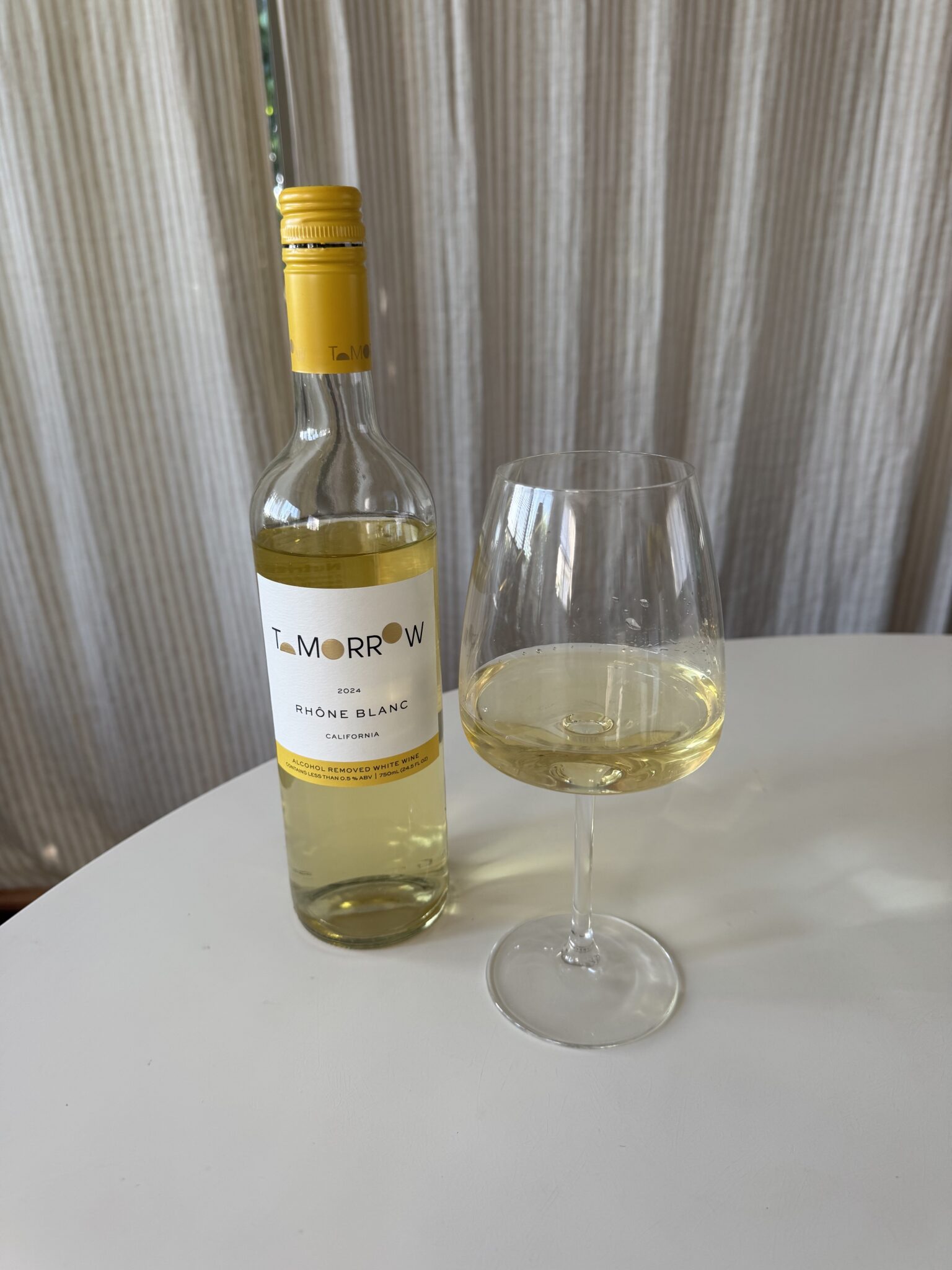 A bottle of Tomorrow Rhone Blanc white wine stands next to a filled wine glass on a round white table with light curtains in the background.