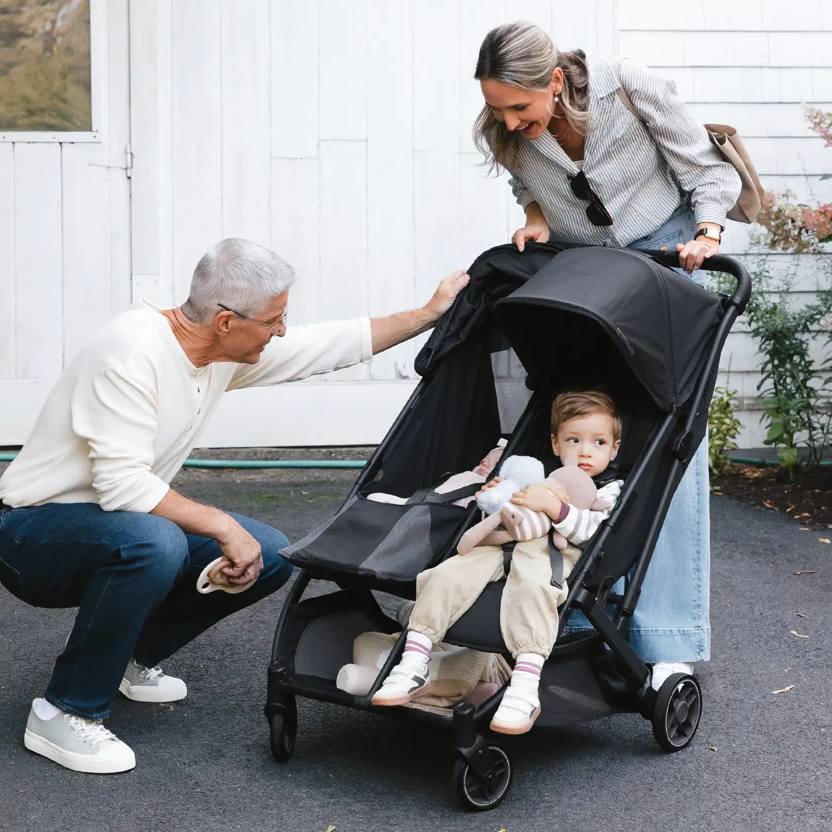 An older man and a woman interact with a toddler seated in a black stroller outside a white building.