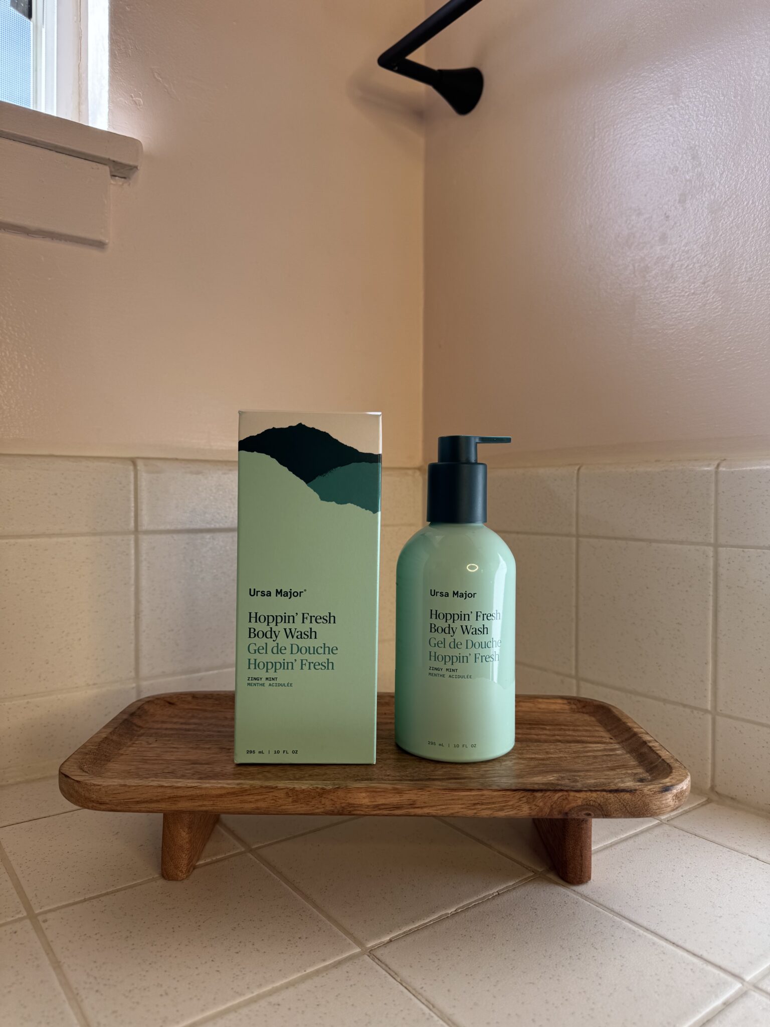 A bottle and box of Ursa Major Hoppin’ Fresh Body Wash are displayed on a wooden tray in a tiled bathroom corner.