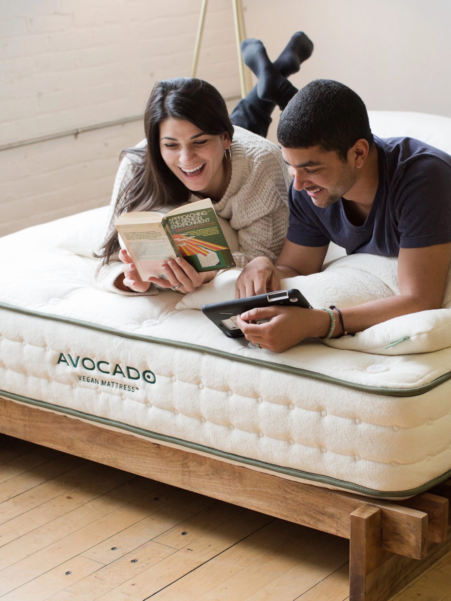 Two people lie on an Avocado vegan mattress; one reads a book while the other looks at a tablet, both smiling and relaxed in a bright room with wooden floors.